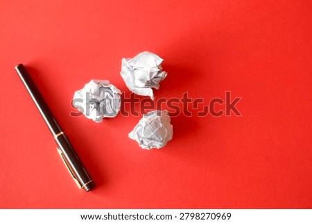 Flat lay top down view of a black executive fountain pen alongside three crumpled white papers isolated on a vibrant red paper background, representing writer's block or discarded ideas.