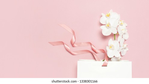 Flat Lay Top View Beautiful White Phalaenopsis Orchid Flowers And White Gift Bag On Pastel Pink Background. Greeting Card With Orchid Tropical Flower. Holiday Birthday Women's Day Floral Card Beauty