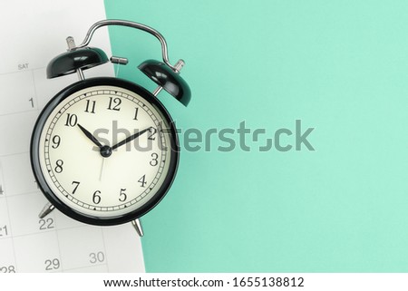 Flat lay or top view  of black alarm clock with white calendar on pastel green background with copy space, schedule for vacation, reminder for business appointment or deadline for business project.