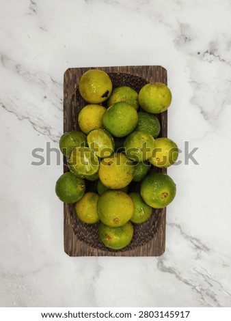 Flat lay top view of fresh whole and halved calamansi limes arranged on a rustic coconut wood tray. Detailed texture of green and yellow peel with visible pores, natural blemishes, and water droplets. Cross section of cut lime shows juicy pulp vesicles, white central pith, and green seeds. Coconut palm wood grain texture visible. Placed on a light stone surface background.