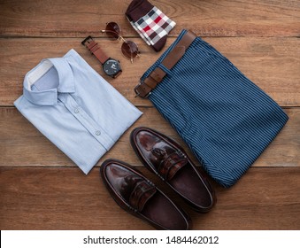 Flat Lay, Top View, Men Fashion Casual Clothing Set On Wooden Background Include Penny Loafer Shoes, Blue Pants, Belt ,watch, Sunglass, Sock And Office Shirt.