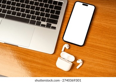 Flat lay top view of modern workspace featuring a silver laptop, blank screen tablet for mockup, and wireless earbuds on a brown wooden desk. Concept for remote work, online learning, and digital noma