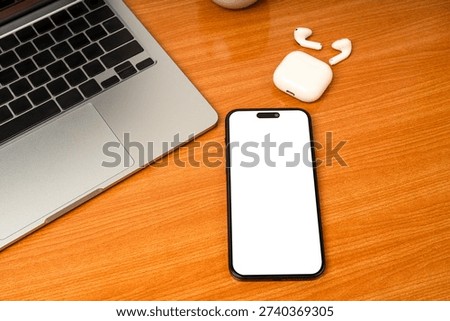 Flat lay top view of modern workspace featuring a silver laptop, blank screen tablet for mockup, and wireless earbuds on a brown wooden desk. Concept for remote work, online learning, and digital noma