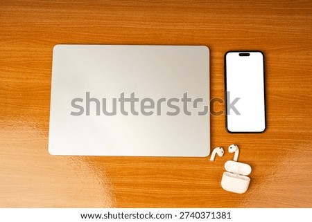 Flat lay top view of modern workspace featuring a silver laptop, blank screen tablet for mockup, and wireless earbuds on a brown wooden desk. Concept for remote work, online learning, and digital noma