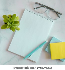 Flat Lay, Top View Office Table White Desk. Workspace With Blank Notepad, Glasses, Pen, Post It Sticker Paper For Notes And Plant In Pot On White Marble Background. Copy Space, Square Instagram Format