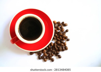 Flat lay top view of a red ceramic espresso cup filled with black coffee placed next to a pile of roasted coffee beans, isolated on a white background