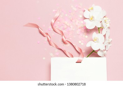 Flat lay top view White Phalaenopsis orchid flowers festive confetti white gift bag on pastel pink background. Greeting card with orchid Tropical flower. Holiday birthday Women's Day floral Card 