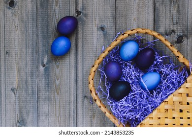 Flat lay top view of a wooden rustic old table on which paper shavings filler in the form of a nest and Easter eggs painted in blue and purple colors are scattered from a wicker golden basket, space. - Powered by Shutterstock