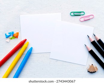 Flat lay of two blank square papers on a light fabric background with colorful crayon pieces, paper clips, pencil shavings, and black pencils. Creative stationery mockup with copy space.