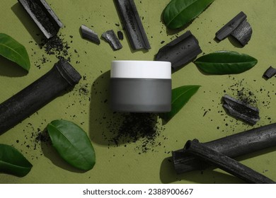 Flat lay of an unlabeled gray cosmetic jar, surrounded by bamboo charcoal and fresh green tea leaves. Mask made from activated bamboo charcoal powder have the effect of cleansing the skin Stock Photo
