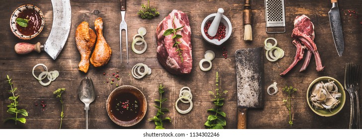 Flat lay of various grill and bbq meat : chicken legs, steaks, lamb ribs with vintage kitchenware kitchen utensils:  Meat Fork and Butcher Cleaver and herbs knife. Sauces and ingredients for grilling,