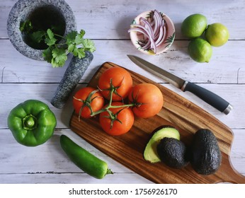 Flat Lay Of Vegetables Including Tomato, Pepper, Jalapeño, Avocado, Lime, Onion And Cilantro Over A Wooden Table And A Mortar And A Pestle. 