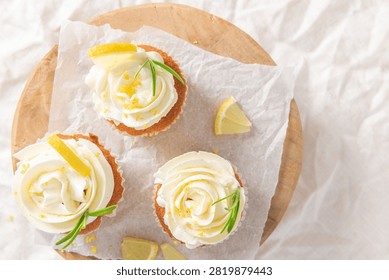 Flat lay view of three delicious homemade lemon cupcakes topped with white cream frosting lemon slices and rosemary sprigs on wooden board