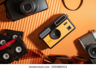 A flat lay of vintage cameras and film reels on an orange background. The focal point is a yellow Kodak camera surrounded by black and silver cameras, and film strips. - Powered by Shutterstock