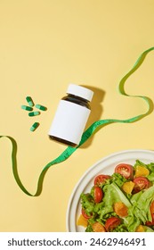 Flat lay weight losing composition on light yellow countertop including a black medicine bottle without label and a dish of vegetable salad. Top view photo with blank space for design