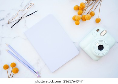 Flat lay of white marble table with stationery, glasses, yellow flowers and photo camera. Top view with copy space. Modern female work place concept
