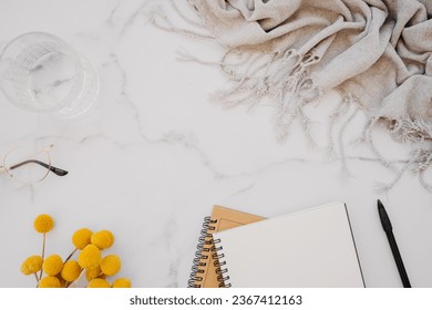 Flat lay of white notebook with copy space for text, glasses and yellow flowers on white marble background. Top view. Minimal home office desk. Work or business concept.