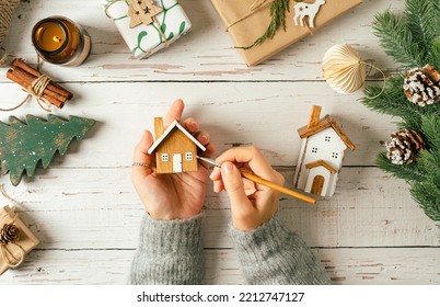 Flat lay of woman hands holding brush, painting homemade wooden house on white background with Christmas natural eco decoration. Top view gifts, candle, fir tree, toy, cinnamon, pine cone, craft. - Powered by Shutterstock