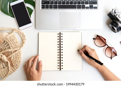 Flat lay of woman hands writing on notebook with travel accessories and laptop on desk. Summer, holiday, work and planning travel concepts. Top view and copy space
