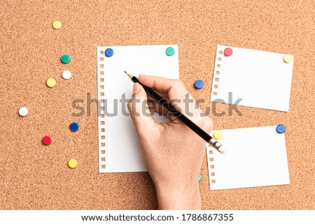 Flat lay of a woman's hand interacting with a pencil in to write on a blank paper attached with colored thumb tacks over a cork board