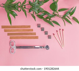 Flat lay of wooden wicks, scissors and wick holders on pink background. Nearby are matches and a branch with green juicy leaves