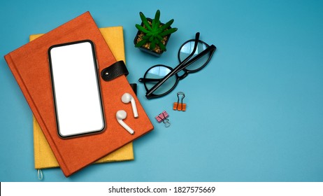 Flat Lay, Workspace Back To School And Education Concept On Blue Table Desk With Blank Screen Phone On Stack Of Book, Earphone And Eyeglass, Green Plant, Top View With Copy Space