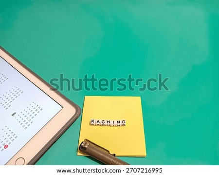 Flat lay of a yellow note reading ‘KaChing’ beside a pen and a digital calendar on a teal background, symbolizing payday, budgeting, financial planning, and modern productivity.