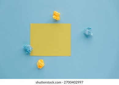 A flat lay of a yellow square note surrounded by crumpled blue and yellow paper on a light blue background. Simple and minimalistic design.