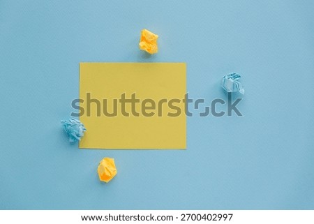A flat lay of a yellow square note surrounded by crumpled blue and yellow paper on a light blue background. Simple and minimalistic design.