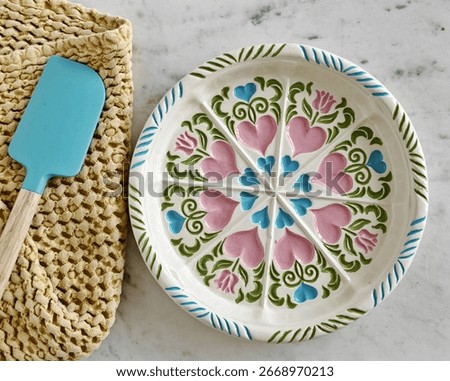 Flat layout of a handmaid’s colourful painted pie plate from the 1980s, including flowers and hearts and pink, blue and green. Also showing complementing blue spatula and yellow towel.