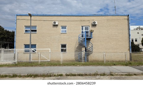 A flat perspective captures a two-storey light brick industrial facility with a blue metal spiral staircase, small windows, and air conditioning units behind a mesh fence