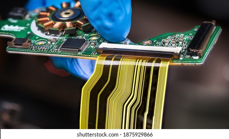 Flat Plastic Strip Flexible Cable On Printed Circuit Board On A Dark Background. Hand In Blue Glove Holding Green PCB With Integrated Motor Stator And Electronic Components As Micro Chip Or Resistors.