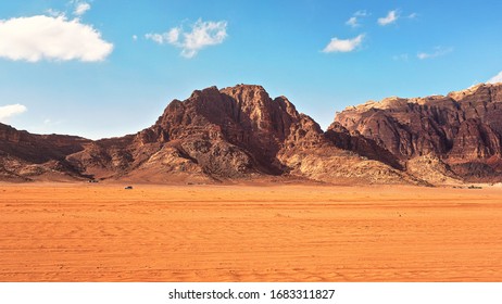 Flat red desert with large mountains in distance, blue sky above, some off road vehicles in background, typical scenery at Wadi Rum, Jordan - Powered by Shutterstock