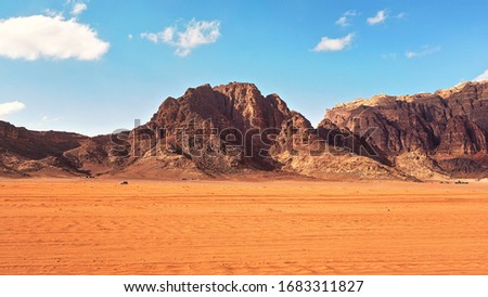 Flat red desert with large mountains in distance, blue sky above, some off road vehicles in background, typical scenery at Wadi Rum, Jordan