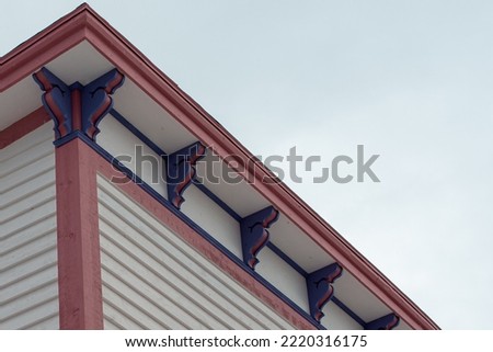 A flat roof corner section of a large white vintage wooden building with decorative pink and purple wood finial trim under the eave. The trim is pink color. The sky is a cloudy blue in color. 