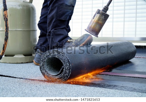 Flat roof installation with propane\
blowtorch during construction works with roofing felt. Heating and\
melting bitumen roofing felt. Roofing felt. Roofer working. Roofer\
working tool.\
Waterproofing