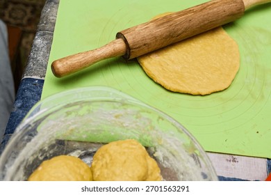 Flatbread dough gets rolled out on green silicone mat using wooden rolling pin in cozy kitchen. Flour dusted dough balls sit in glass bowl, adding rustic charm to homemade bread-making scene.