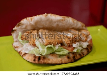 Flatbread sandwich filled with breaded chicken cutlet, lettuce and tomato, served on green plate, close view of crispy coating and soft bread, cafe lunch or takeaway meal.