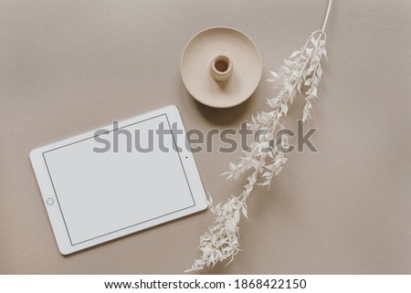 Flatlay of blank screen tablet pad, white floral branch, decorations on neutral pastel beige background. Home office desk workspace. Business, work template. Flat lay, top view.