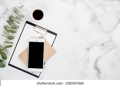 Flatlay Business Home Office Desk With Office Supplies And Smartphone. Top View With Copy Space. Mobile Phone, Eucalyptus Branch, Clipboard, Paper Notebook And Cup Of Coffee On Marble Table.