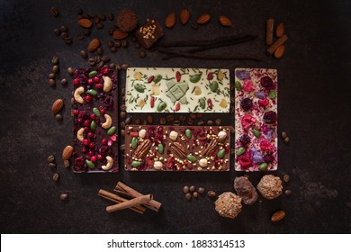 A flat-lay composition of beautiful hand-made dark and white chocolate bars containing nuts and berries, chocolate candies, nuts, cinnamon sticks and coffee beans lying on a dark background