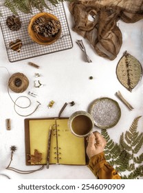 A flat-lay composition featuring a hand holding a cup of coffee, an open notebook with a wooden pencil  on a white textured surface. Perfect for themes of journaling, autumn, or creative workspace