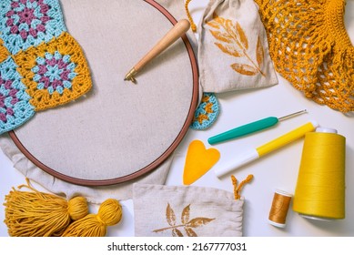 Flatlay Of Embroidery Hoop With Blank Fabric And Punch Needle Inserted In, Crochet Hook, Tassels, Heart, Spool Of Thread, Marker And Crochet Patterns On White Surface. 