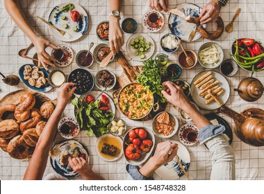 Flat-lay Of Family Sitting At Table With Turkish Breakfast With Pastries, Vegetables, Greens, Spreads, Cheeses, Eggs, Jams And Tea In Tulip Glasses And Copper Teapots Over Chekered Linen Tablecloth