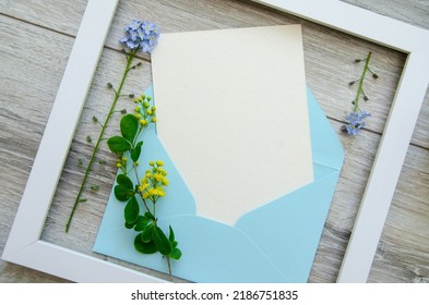 Flatlay Gray Natural Wooden Background With  Square White Wooden Frame And  Blue Envelope White Postcard Copyspace, Small Blue And Yellow Flowers And Green  Leaves On A Branch