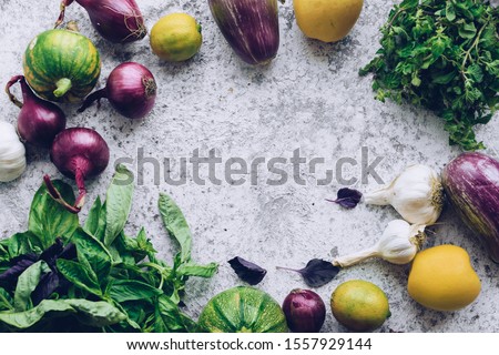 Flat-lay of green and purple raw vegetables over grey background, food frame. Local produce for healthy cooking. Eggplans, red onion, mint, garlic and basil. Clean eating. Top view. Copy space.