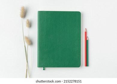 Flatlay Of Home Office Desk Table. Green Notebook With Green And Red Pencils And Dry Flower On White Background