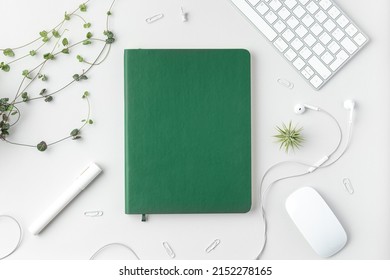 Flatlay Of Home Office Desk Table. Top View Of Workspace With Green Notebook, Keyboard, Mouse, Marker, Headphones, Pins And Plants On White Background.