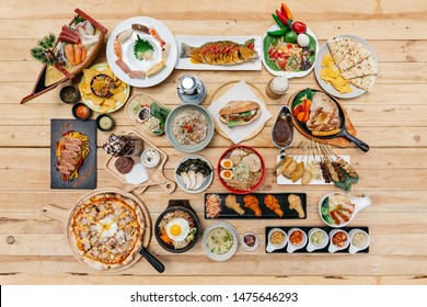 Flatlay Of International Foods On Wooden Table: Sushi, Sashimi, Nachos, Spaghetti, Naan, Sandwich, Noodle, Steak, Ramen. Yakitori, Pizza, Bibimbap, Fried Chicken Wings, Wonton Soup And Brownie Frappe.