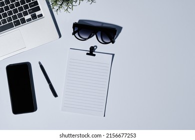 Flatlay of keyboard of laptop, stylish sunglasses, smartphone, pen and blank paper with clip with copyspace for your text on the right